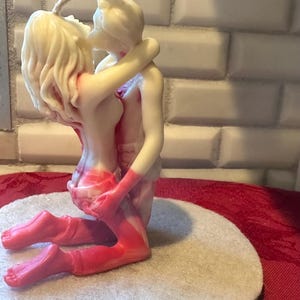K&ouml;nnte beinhalten: Eine cremefarbene Kerze, die zwei sich umarmende Figuren darstellt. Die Figuren sind in einer leidenschaftlichen Umarmung verschlungen, mit roten Akzenten an Beinen und Torso. Die Kerze steht auf einer runden, cremefarbenen Filzbasis.
