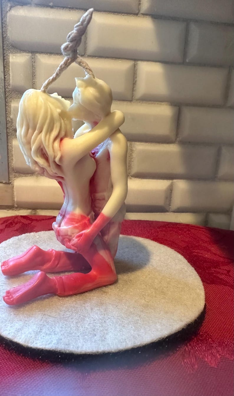 K&ouml;nnte beinhalten: Eine cremefarbene Kerze, die zwei sich umarmende Figuren darstellt. Die Figuren sind in einer leidenschaftlichen Umarmung verschlungen, mit roten Akzenten an Beinen und Torso. Die Kerze steht auf einer runden, cremefarbenen Filzbasis.