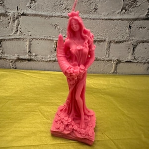 May include: A pink candle in the shape of a woman with long flowing hair, a crown, and a horn. The candle is holding a bouquet of flowers and is standing on a base decorated with flowers. The background is a white brick wall.