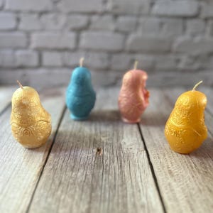 May include: Four pear-shaped candles in gold, blue, pink, and yellow are arranged on a wooden surface. The candles have a textured surface and a wick at the top. The background is a blurred white brick wall.