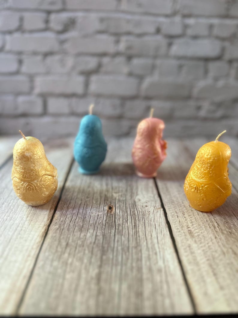 May include: Four pear-shaped candles in gold, blue, pink, and yellow are arranged on a wooden surface. The candles have a textured surface and a wick at the top. The background is a blurred white brick wall.