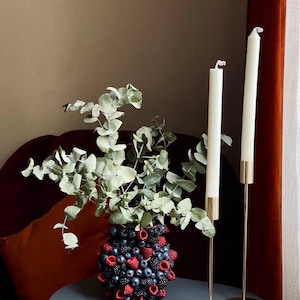 May include: A decorative arrangement featuring a vase adorned with red and blue berries, topped with green foliage. Two white candles in gold-toned holders stand nearby. The items are arranged on a gray table, creating a stylish home decor scene.