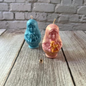 May include: Two Matryoshka doll candles. One is light blue, the other is pink. Both dolls have a face and are holding a toy horse. The candles are on a wooden surface with a brick background.