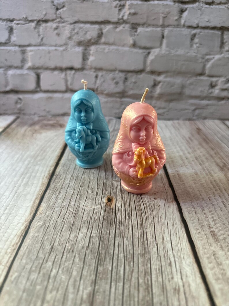 May include: Two Matryoshka doll candles. One is light blue, the other is pink. Both dolls have a face and are holding a toy horse. The candles are on a wooden surface with a brick background.