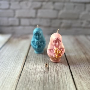 May include: Two decorative candles in the shape of a woman, one light blue and the other pink. The candles have a detailed design and a wick at the top. They are placed on a wooden surface with a brick wall background.