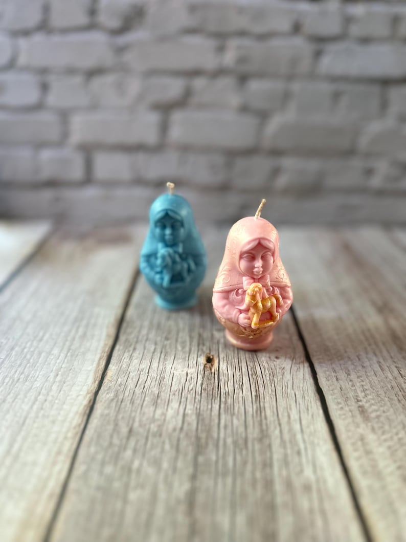 May include: Two decorative candles in the shape of a woman, one light blue and the other pink. The candles have a detailed design and a wick at the top. They are placed on a wooden surface with a brick wall background.