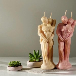 Pode incluir: Duas velas decorativas em forma de figuras abraçadas, uma bege clara e outra rosa. Uma pequena planta suculenta num vaso rosa claro está em primeiro plano. O fundo é de uma cor neutra.