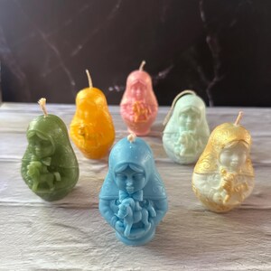 May include: A collection of six pear-shaped candles in various colors, including green, orange, pink, light blue, and gold. Each candle features a detailed relief of a religious figure. The candles have wicks at the top and are arranged on a light-colored wooden surface.