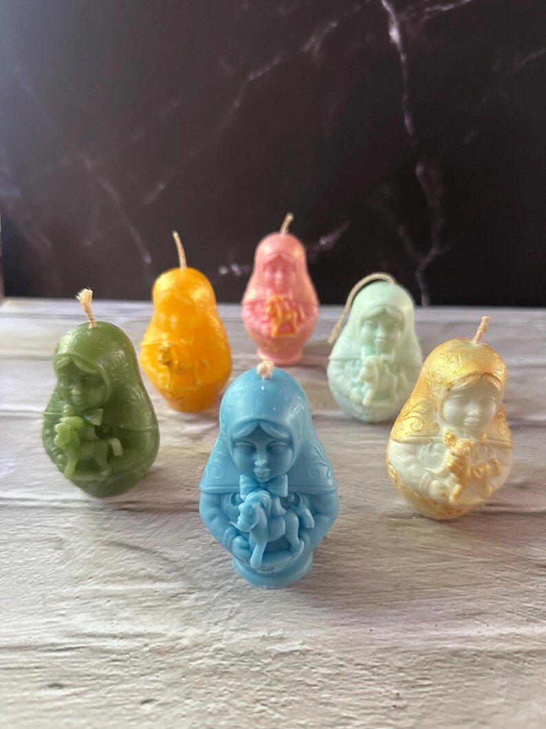 May include: A collection of six pear-shaped candles in various colors, including green, orange, pink, light blue, and gold. Each candle features a detailed relief of a religious figure. The candles have wicks at the top and are arranged on a light-colored wooden surface.