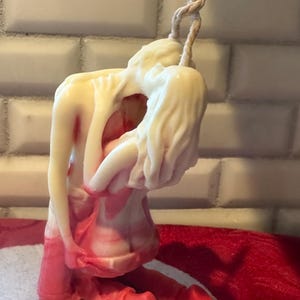K&ouml;nnte beinhalten: Eine Kerzenskulptur, die zwei sich umarmende Figuren darstellt. Die Figuren sind &uuml;berwiegend cremefarben mit roten Akzenten, vor einem wei&szlig;en, ziegelartigen Hintergrund. Ein verdrehtes, seilartiges Element erstreckt sich von der Oberseite der Skulptur.