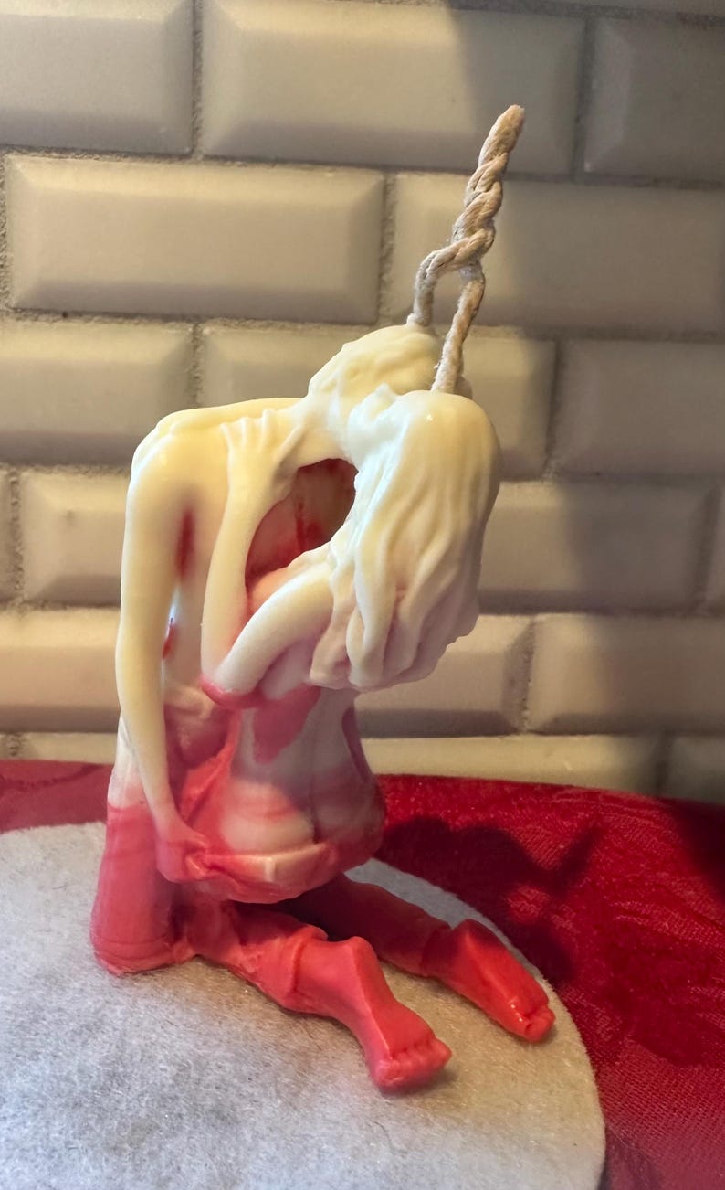 K&ouml;nnte beinhalten: Eine Kerzenskulptur, die zwei sich umarmende Figuren darstellt. Die Figuren sind &uuml;berwiegend cremefarben mit roten Akzenten, vor einem wei&szlig;en, ziegelartigen Hintergrund. Ein verdrehtes, seilartiges Element erstreckt sich von der Oberseite der Skulptur.