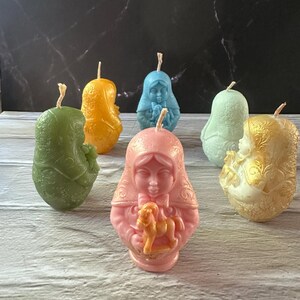 May include: A collection of six decorative candles in various colors, including green, orange, blue, light green, pink, and gold. Each candle is shaped like a person with a detailed design. The candles are arranged on a light-colored surface.