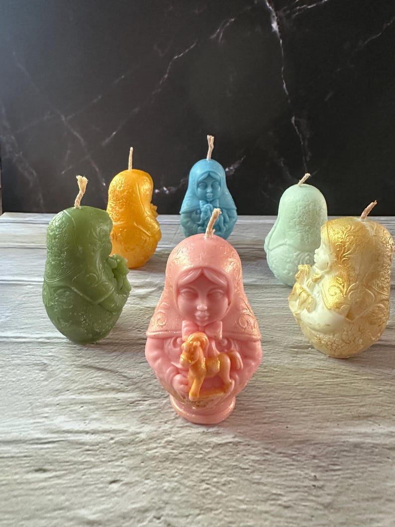 May include: A collection of six decorative candles in various colors, including green, orange, blue, light green, pink, and gold. Each candle is shaped like a person with a detailed design. The candles are arranged on a light-colored surface.