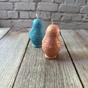 May include: Two decorative candles shaped like pears. One is light blue, the other is pink with gold accents. Both have a detailed floral design on the lower half and a wick at the top. The candles are on a wooden surface.