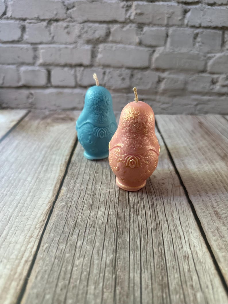 May include: Two decorative candles shaped like pears. One is light blue, the other is pink with gold accents. Both have a detailed floral design on the lower half and a wick at the top. The candles are on a wooden surface.