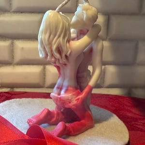 Könnte beinhalten: Eine Kerze, die zwei sich umarmende Figuren zeigt, mit einem rot-weißen Farbschema. Die Figuren sind miteinander verschlungen, mit einem Docht, der sich von oben erstreckt. Die Kerze steht auf einem Filzherz und rotem Band.
