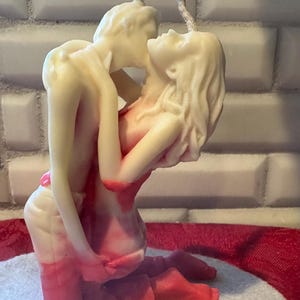 K&ouml;nnte beinhalten: Eine cremefarbene Kerze, die ein sich umarmendes Paar darstellt, mit roten Akzenten auf ihren K&ouml;rpern. Die Figuren sind in einer leidenschaftlichen Pose verschlungen, mit einem gedrehten Docht, der sich von oben erstreckt. Die Kerze steht vor einem neutralen Hintergrund.