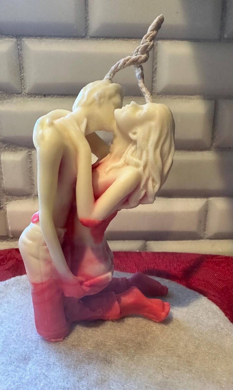 K&ouml;nnte beinhalten: Eine cremefarbene Kerze, die ein sich umarmendes Paar darstellt, mit roten Akzenten auf ihren K&ouml;rpern. Die Figuren sind in einer leidenschaftlichen Pose verschlungen, mit einem gedrehten Docht, der sich von oben erstreckt. Die Kerze steht vor einem neutralen Hintergrund.