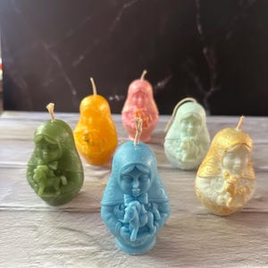 May include: A collection of six colorful, pear-shaped candles. The candles are in shades of green, orange, pink, light green, blue, and gold. Each candle features a detailed design of a woman holding a child. The candles have a rustic, handmade appearance.