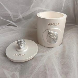 Op de afbeelding: Een crèmekleurige decoratieve pot met een deksel. De pot heeft een zilveren reliëf van een klassieke buste en het woord "APOLLO". Het deksel heeft een bijpassende buste en het woord "APOLLO". De pot is cilindrisch en staat op een witte ondergrond.
