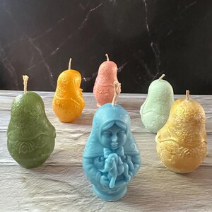 May include: A collection of colorful, decorative candles in various shapes. The candles include a blue figure of a woman holding a baby, and pear-shaped candles in green, orange, pink, and gold. Each candle has a wick.