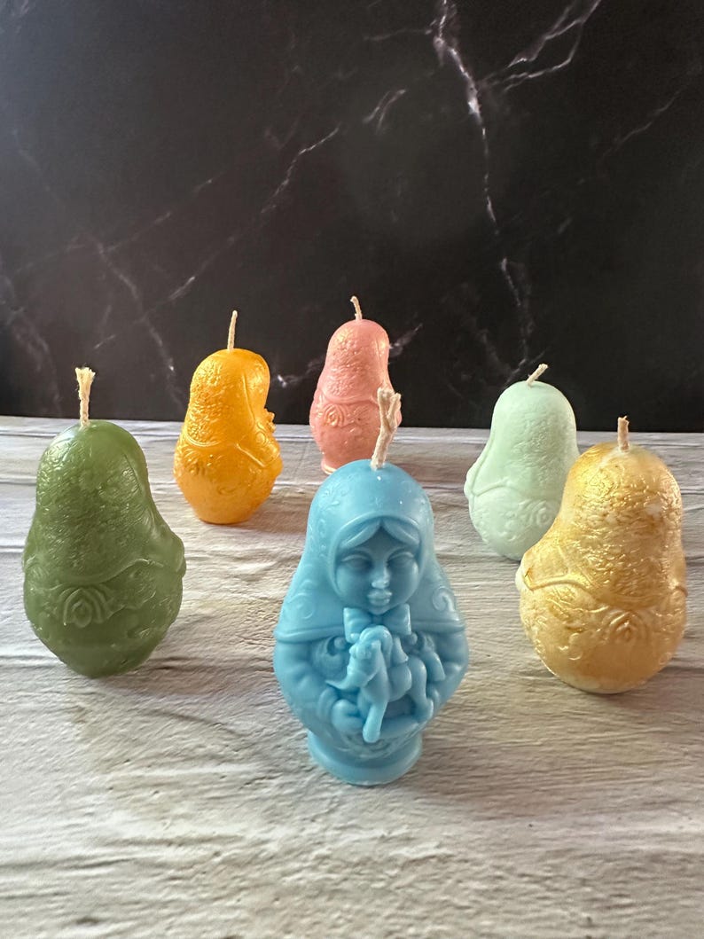 May include: A collection of colorful, decorative candles in various shapes. The candles include a blue figure of a woman holding a baby, and pear-shaped candles in green, orange, pink, and gold. Each candle has a wick.