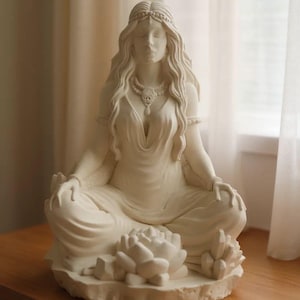 Puede incluir: Estatua de color crema de una mujer sentada en pose meditativa. La estatua presenta cabello largo y ondulado, un collar detallado y una base de flor de loto. La mujer lleva un vestido fluido y tiene los ojos cerrados.