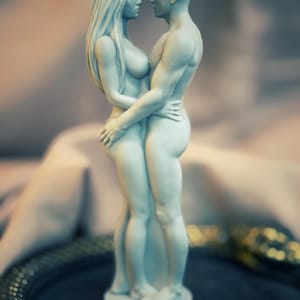 May include: A light blue statue of a man and woman embracing. The figures are nude, with the woman's long hair flowing down her back. The statue is on a round base.