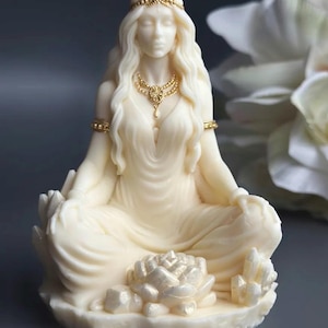 May include: Ivory-colored statue of a seated figure in a meditative pose. The figure wears gold jewelry, including a necklace and headpiece. A lotus flower and crystals are at the base. The statue has long, flowing hair and is draped in a flowing gown.