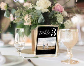 Puede incluir: Un cartel de número de mesa con marco dorado y fondo negro. El número "3" está en blanco y la palabra "mesa" está en blanco en cursiva. Una foto de una pareja en un campo está en el fondo. El texto "Olivia & Graham 27.05.2023" está en la parte inferior del cartel.