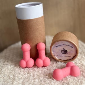 Könnte beinhalten: Rosa Wachs-Schmelzstücke in Form eines Phallus liegen auf einer weißen, flauschigen Oberfläche. Zwei zylindrische Pappbehälter sind ebenfalls vorhanden, einer offen und einer geschlossen, mit Text auf dem Etikett. Die Schmelzstücke sind leuchtend rosa.