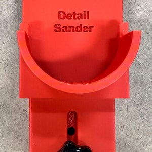 May include: A bright red detail sander with the words "Detail Sander" printed on the front. The tool has a curved sanding surface and a black star-shaped knob for adjustments. It is designed for detailed woodworking projects.