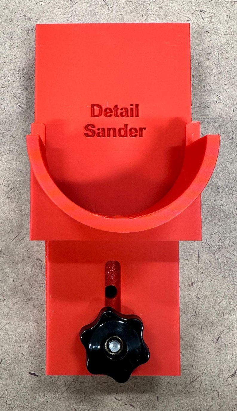 May include: A bright red detail sander with the words "Detail Sander" printed on the front. The tool has a curved sanding surface and a black star-shaped knob for adjustments. It is designed for detailed woodworking projects.