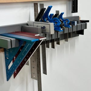 May include: A collection of woodworking tools, including blue speed squares, metal rulers, and a gray Empire square, mounted on a gray rack against a white wall. The tools are organized for easy access.