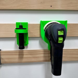 Könnte beinhalten: Zwei grüne, wandmontierte Werkzeughalter mit einem schwarzen Elektrowerkzeug und einem schwarzen und grünen Elektrowerkzeug. Die Elektrowerkzeuge sind zum Schleifen und Polieren.