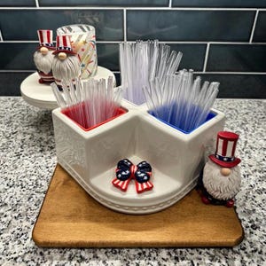May include: A white ceramic caddy with red and blue compartments filled with clear straws. Decorative gnomes in patriotic hats and a bow tie accent the caddy. The caddy sits on a wooden tray, perfect for a Fourth of July celebration.