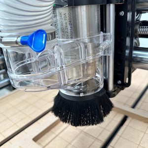 May include: Close-up of a CNC machine with a clear plastic dust shoe and a black brush head. The machine is cutting into a light-colored wooden surface. The text "EDLINE CNC" is visible on the machine.