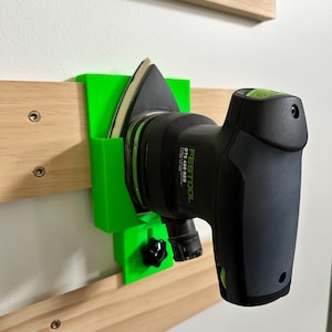 Könnte beinhalten: Eine grüne, 3D-gedruckte Wandhalterung für eine Festool DTS 400 REQ Schleifmaschine. Die Schleifmaschine ist schwarz mit grünen Akzenten und hängt an der Halterung.