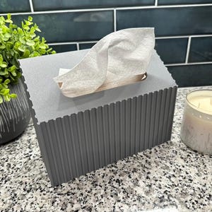 May include: A gray tissue box with a textured, ribbed design. White tissues are pulled from the top opening. A small potted plant and a candle in a glass jar are also visible.