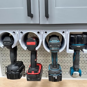 May include: A gray, 3D-printed drill holder with four circular slots, mounted under a gray cabinet. Four black and red power drills are held in place. The holder is attached to a pegboard.