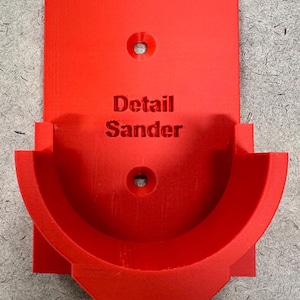 May include: A bright red 3D-printed detail sander holder. The holder features a semi-circular cradle and a rectangular backplate with two mounting holes. The words "Detail Sander" are embossed on the front.