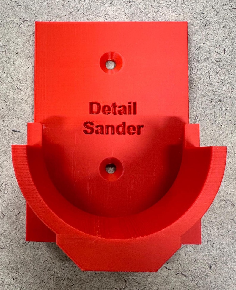 May include: A bright red 3D-printed detail sander holder. The holder features a semi-circular cradle and a rectangular backplate with two mounting holes. The words "Detail Sander" are embossed on the front.