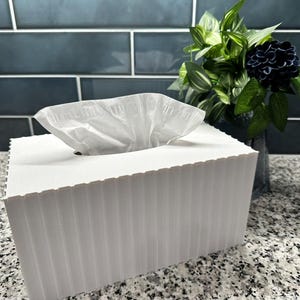 May include: A white tissue box with a ridged design, filled with white tissues. The box is on a speckled countertop, with a dark blue tile backsplash and a vase of greenery in the background.
