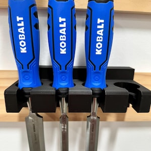 Könnte beinhalten: Drei Stecheisen mit blauen Griffen und dem weißen Markennamen "KOBALT". Sie werden in einem schwarzen Halter auf einem Holzregal gehalten. Die Stecheisenklingen sind silberfarben.