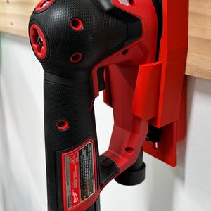 May include: A red and black power tool, mounted on a red holder, attached to a wooden plank. The tool has a textured black grip and red accents. The holder is mounted on a white wall. The tool is a Milwaukee Detail Sander.