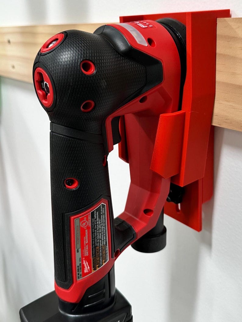 May include: A red and black power tool, mounted on a red holder, attached to a wooden plank. The tool has a textured black grip and red accents. The holder is mounted on a white wall. The tool is a Milwaukee Detail Sander.
