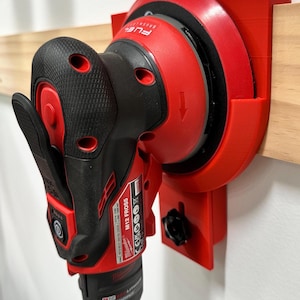 May include: A red and black M12 FROS5 Milwaukee power tool is mounted on a red bracket attached to a wooden surface. The tool has a black handle and a red sanding pad. The bracket is designed to hold the tool securely.