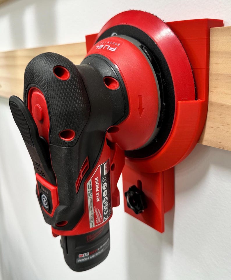 May include: A red and black M12 FROS5 Milwaukee power tool is mounted on a red bracket attached to a wooden surface. The tool has a black handle and a red sanding pad. The bracket is designed to hold the tool securely.