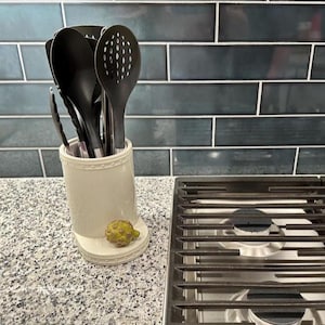 May include: A collection of black kitchen utensils, including a slotted spoon, tongs, and solid spoons, are stored in a cream-colored ceramic utensil holder. A small, green artichoke-shaped decorative item sits on the base. The background features a blue tile backsplash.