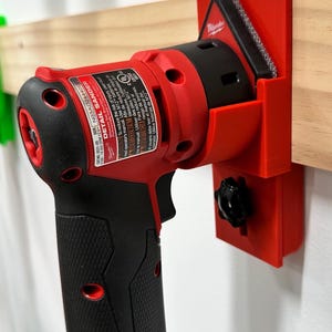 May include: A red and black Detail Sander power tool with a black handle is mounted on a red holder. The tool is attached to a wooden beam. The tool has the text "DETAIL SANDER" and "M12" printed on it.